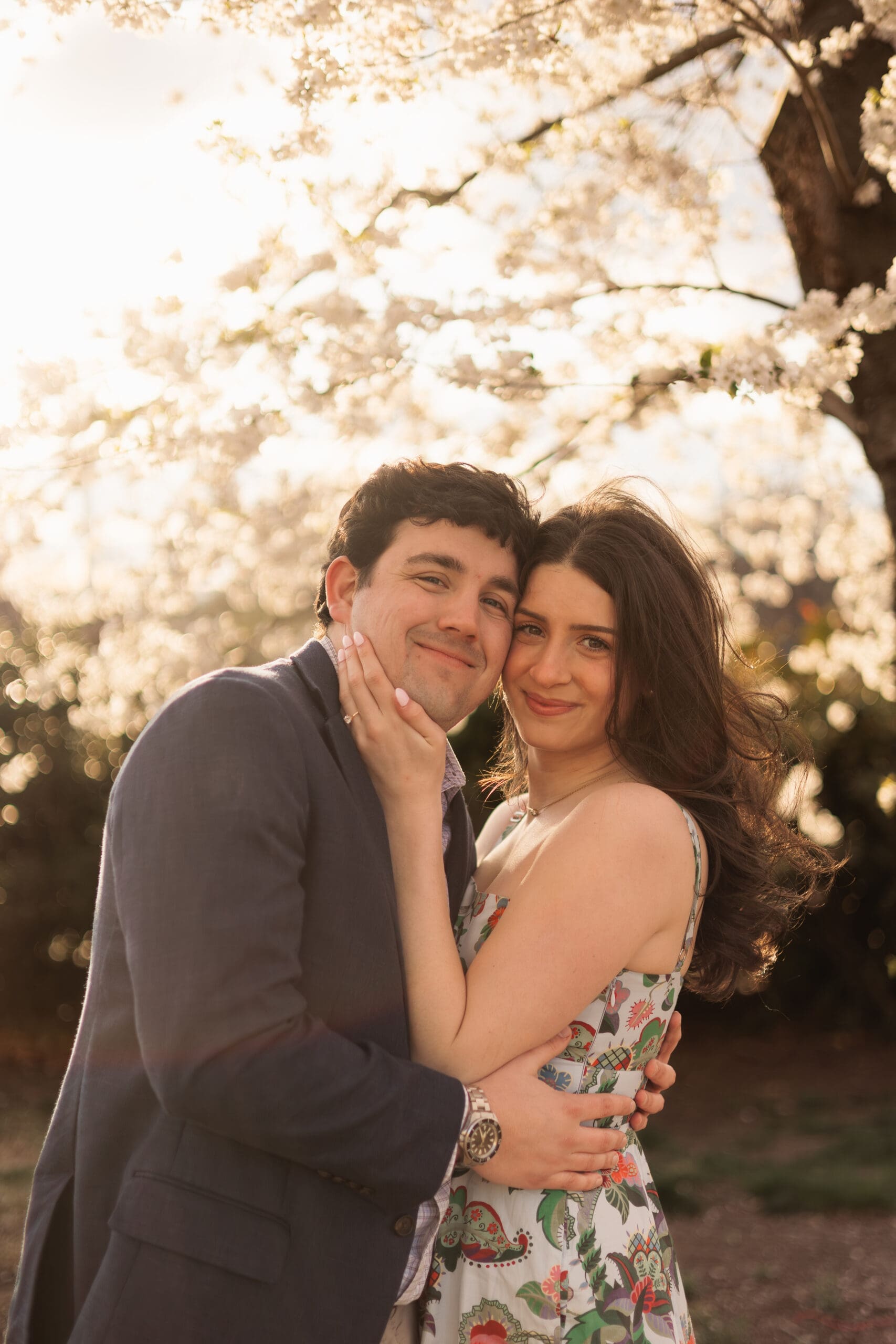 couple smiles for an engagement photo in front of the DC cherry blossoms during a spring, golden hour photoshoot