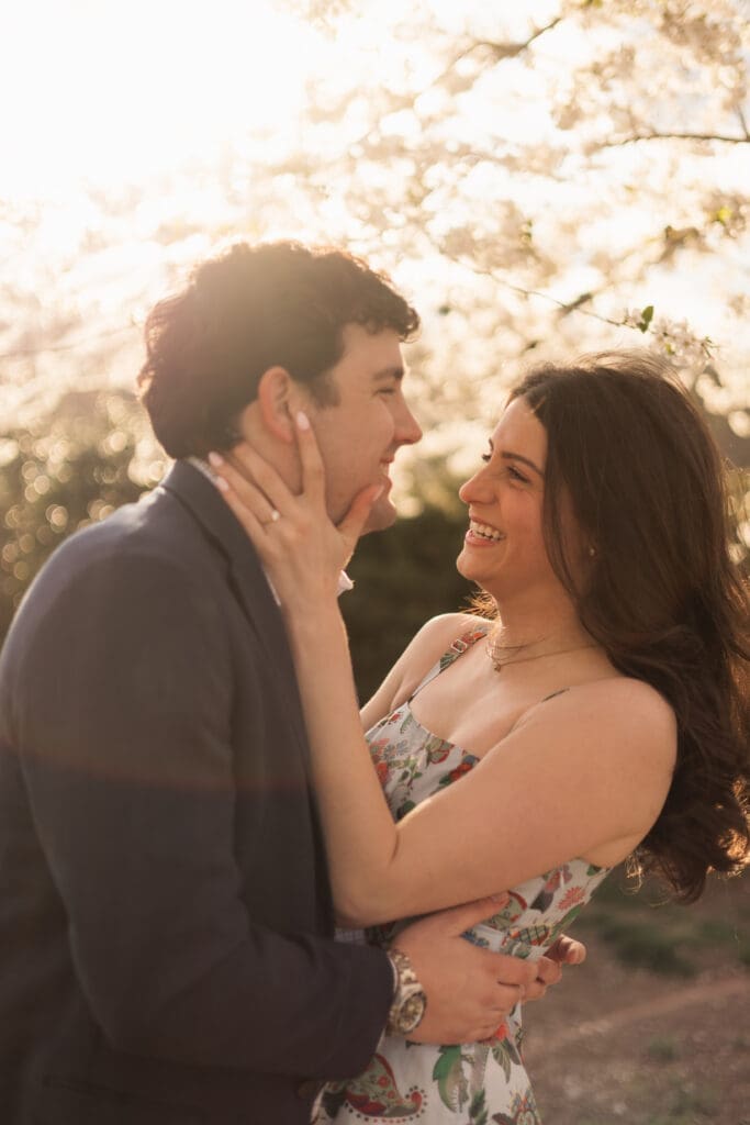 Arlington couples smiles at each other while surrounded by DC cherry blossoms as golden light glows behind them